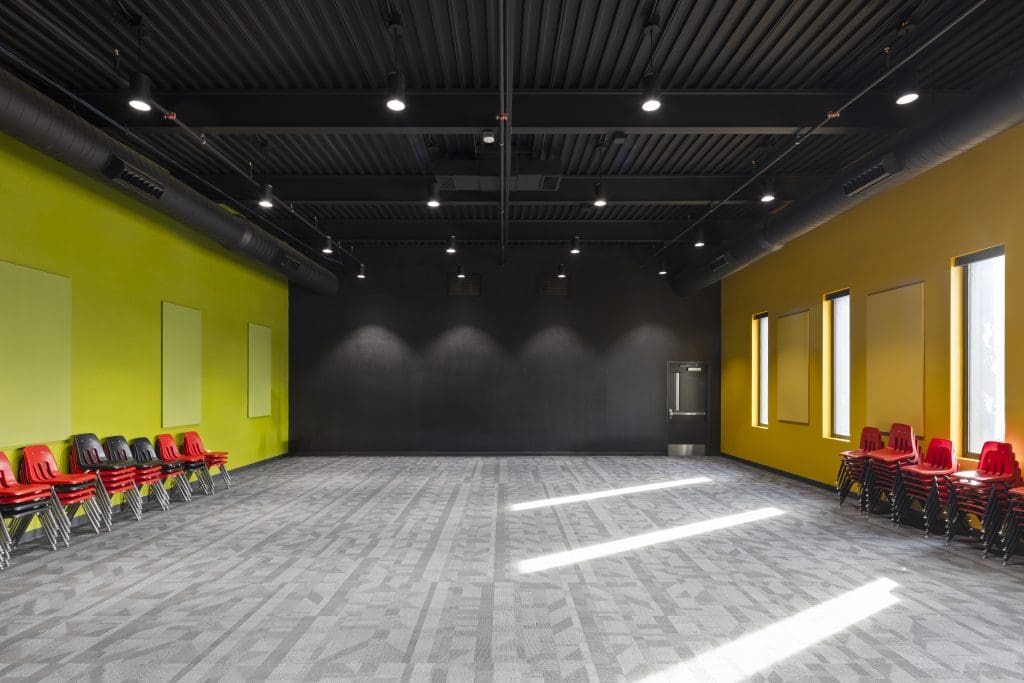 Empty modern room with gray carpet, yellow and black walls, and ceiling lights—part of Faith Christian Church's new building addition. Stacks of red and black chairs line the sides as sunlight streams through windows on the right.
